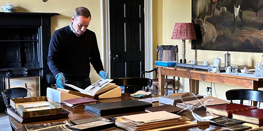 Man wearing a blue jumper and wearing blue gloves looking at books and archive material in what looks like a drawing room.