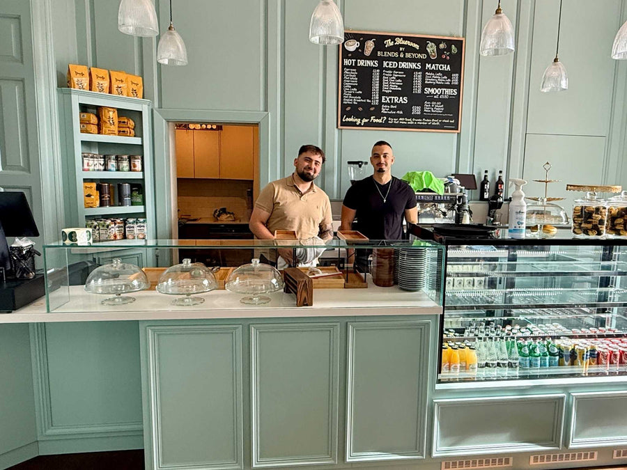 Two men standing beyond a coffee shop counter in a blue period-style room
