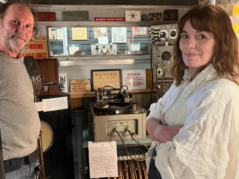 Man with a grey r-shirt standing with a woman with her arms folded and smiling wearing a white shirt in front of retro audio equipment