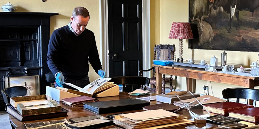 Man wearing a blue jumper and wearing blue gloves looking at books and archive material in what looks like a drawing room.