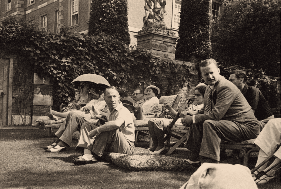 Sepia image from the 1920s of a group of men and women seated in chairs and sitting on the floor in conversation with some of a stately home shown un the background