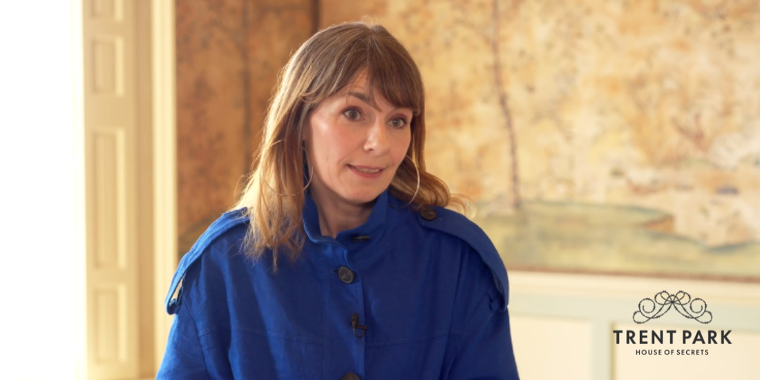 Colour photograph of a white woman with mid brown hair, wearing a colbalt blue jack, standing in a room with sepia toned Chinese wallpaper