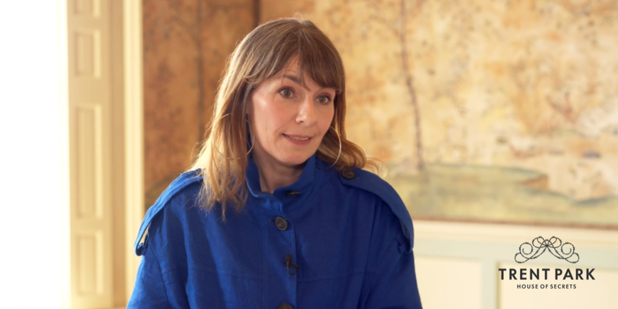 Colour photograph of a white woman with mid brown hair, wearing a colbalt blue jack, standing in a room with sepia toned Chinese wallpaper