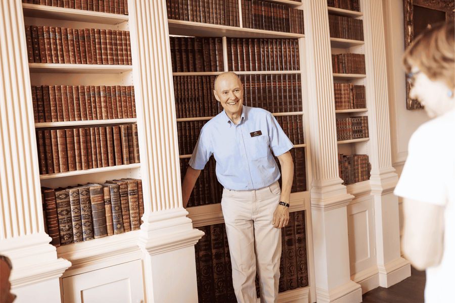 A smiling older man wearing a blue shirt and wearing cream trousers holds what looks like a hidden bookcase door